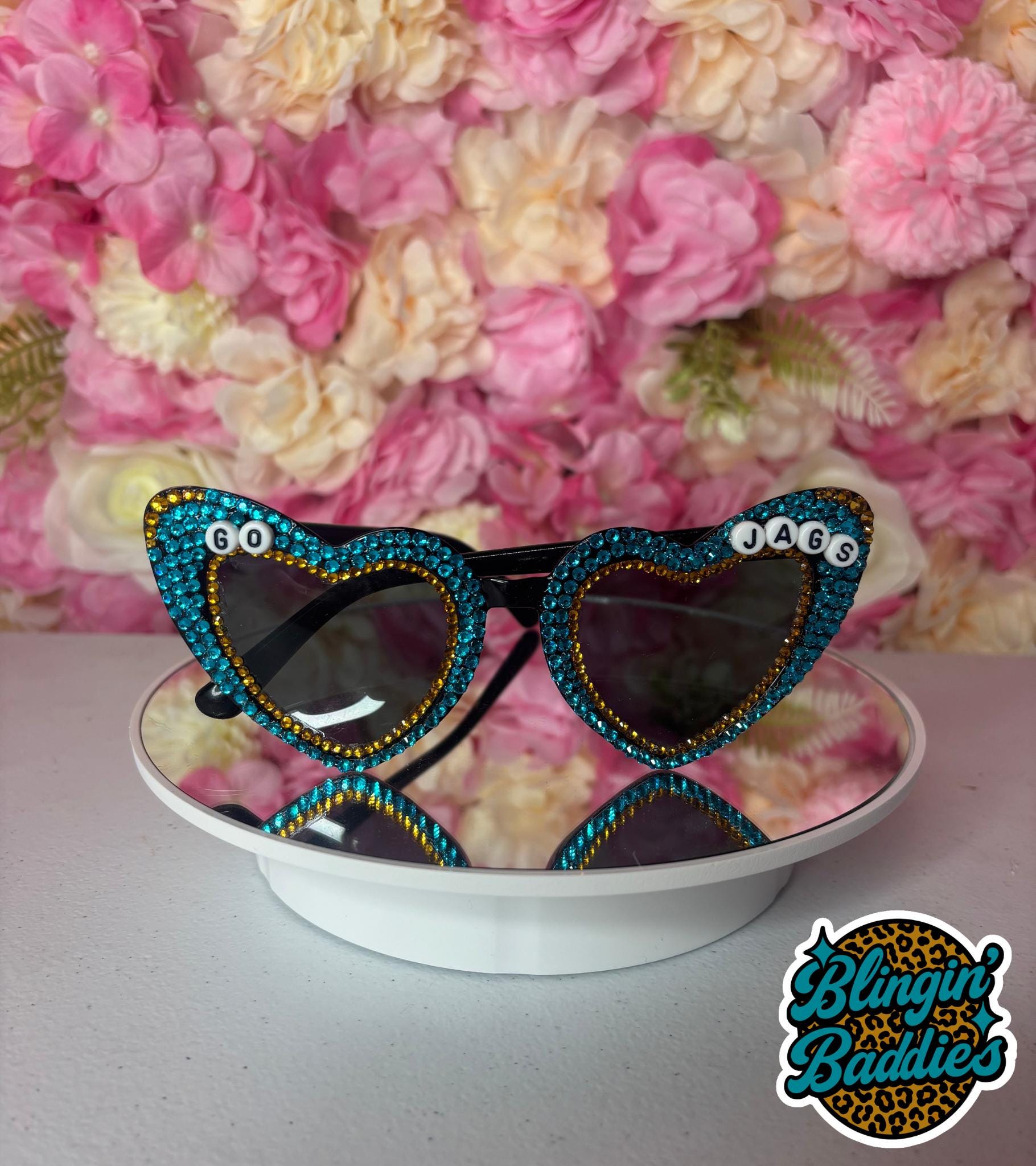 Front view of GO JAGS teal rhinestone heart sunglasses with letter beads against pink floral wall